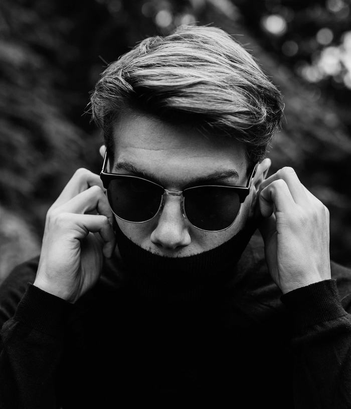 Black & white photograph of a man wearing stylish sunglasses with a turtleneck sweater pulled up to his mouth