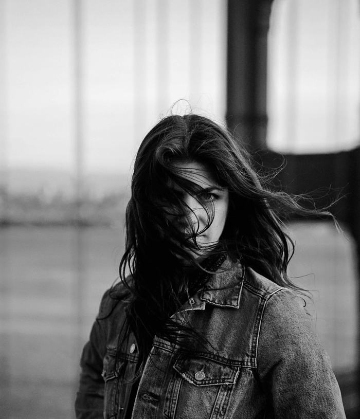 Black & white photograph of a woman, her hair tussled by the ocean wind, standing in front of the Golden Gate Bridge in soft focus