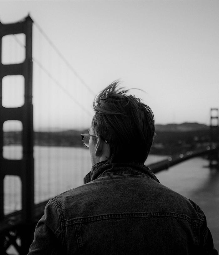 Black & white photo of a man, standing back to camera, gazing out at the Golden Gate Bridge in soft focus