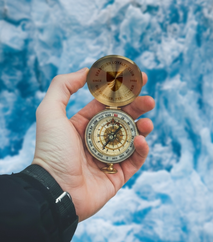 A hand holds an open brass compass against bright blue glacial ice. The compass lid is engraved with a shield logo and the text "SEABOURN LOYALTY CLUB, SINCE 1986, GO FARTHER. FEEL CLOSER."