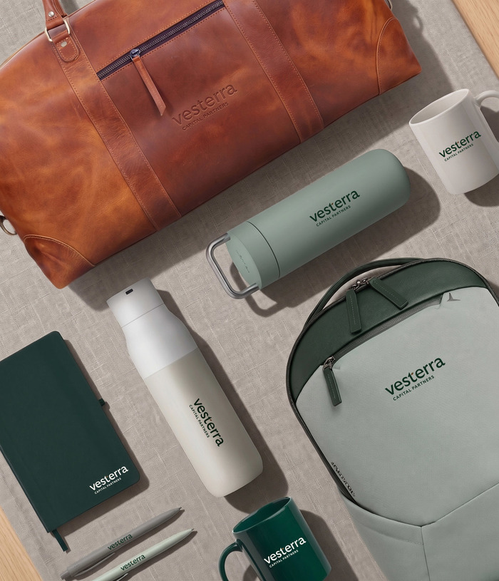Vesterra-branded water bottles, a leather bag, a notebook, mugs and pens laying flat on a tabletop