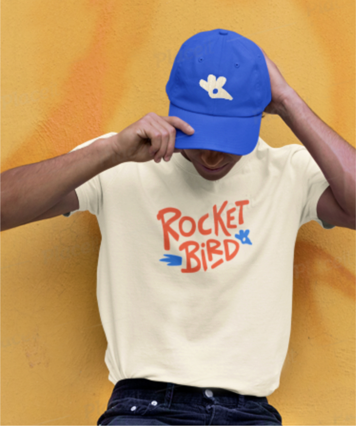 Person posing against yellow wall wearing white rocketbird sandwich and blue rocketbird hat